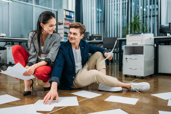 Jeunes partenaires d'affaires heureux faisant de la paperasse tout en étant assis sur le sol au bureau — Photo de stock