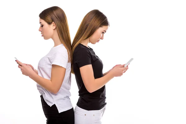 Вид сбоку близнецов, стоящих спиной к спине и использующих смартфоны, изолированные на белом фоне — стоковое фото