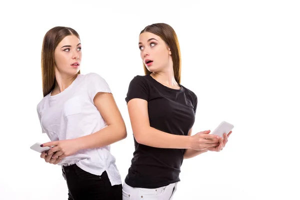 Portrait of shocked twins with smartphones looking at each other isolated on white — Stock Photo