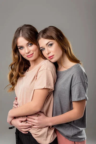 Portrait of beautiful twin sisters hugging each other and looking at camera isolated on grey — Stock Photo