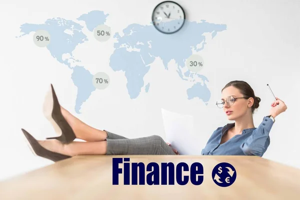 Businesswoman reading documents and sitting with legs on table with finance inscription and world map — Stock Photo