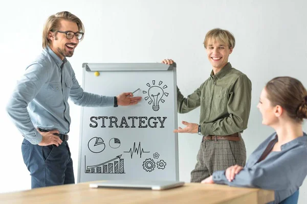 Hombres de negocios riendo mostrando la inscripción de la estrategia y signos de negocio en flipchat a la mujer de negocios - foto de stock
