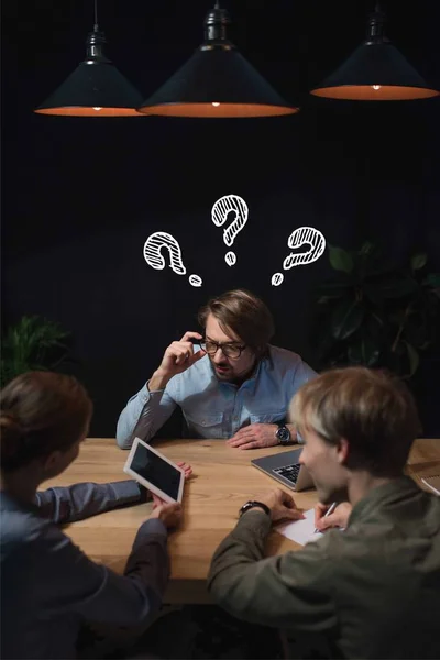 Businesswoman showing something on tablet to her partners with question marks over head and sitting at table — Stock Photo