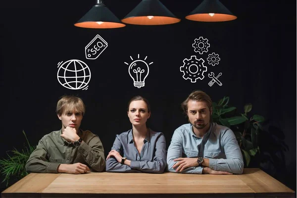Empresária e empresários sentados à mesa em quarto escuro, com ícones de negócios — Fotografia de Stock