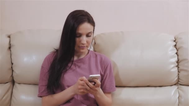 Ung brunett sitter på lädersoffa hemma och använder Smat telefon — Stockvideo