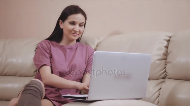 Jeune fille brune dans une chaussettes chaudes bas, pose sur le canapé en cuir à la maison et de travail sur ordinateur portable, achats en ligne avec des cartes en plastique — Video