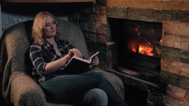 Όμορφη νεαρή γυναίκα διαβάζοντας το βιβλίο κάθεται με τζάκι σε ένα εξοχικό σπίτι. Διακοπές και ιδέα του τρόπου ζωής — Αρχείο Βίντεο
