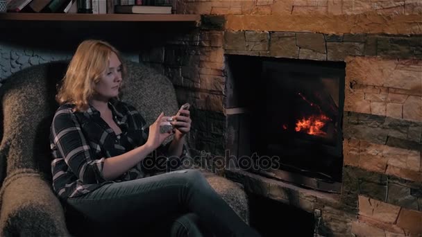 Hermosa mujer usando el teléfono inteligente sentado junto a la chimenea en casa, vacaciones y estilo de vida Concepto — Vídeo de stock