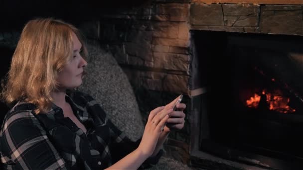 美丽的女人使用智能手机的壁炉坐在家里，节日和生活方式的概念 — 图库视频影像