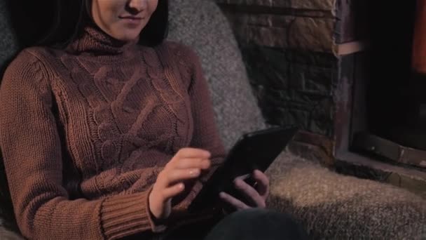 Mujer joven usando tableta digital sentada junto a la chimenea en casa, primer plano — Vídeo de stock