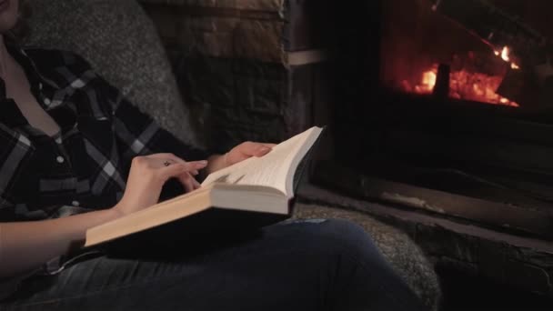 Vacker ung kvinna läsa bok sitter vid brasan i ett hus på landet. Semester- och livsstilskoncept — Stockvideo
