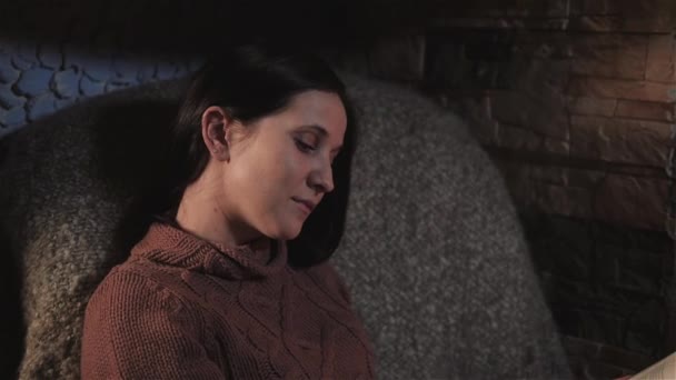 Hermosa joven leyendo libro sentado junto a la chimenea en una casa de campo. Primer plano SHot, vacaciones y estilo de vida Concepto — Vídeo de stock