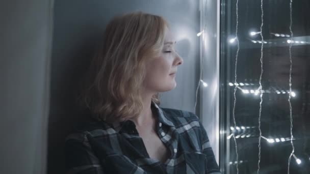 Mooi aantrekkelijke vrouw zittend op de vensterbank versierd met slingers. Concept van Nieuwjaar, Kerstmis, vakantie — Stockvideo