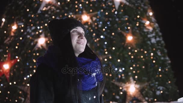 Fiatal, vonzó nő csillogó karácsonyfa előtt áll. Nő a meleg ruhát külső éjjel keresi a kamera, és mosolyogva. — Stock videók