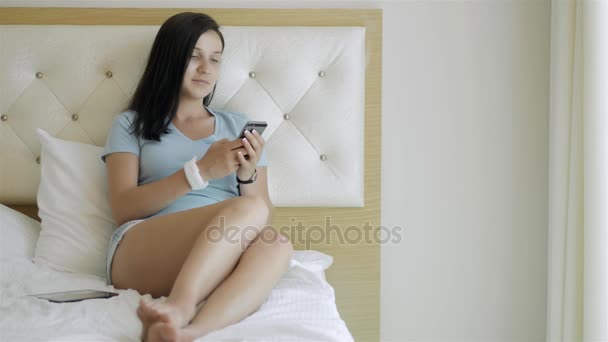 Молода брюнетка дівчина використовує смартфон лежить на ліжку вранці — стокове відео