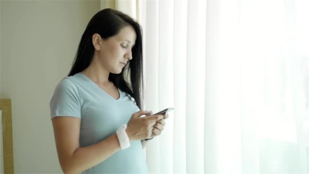 Портрет молодой женщины с помощью смартфона. Кавказская модель стоит у окна и пользуется смартфоном — стоковое видео