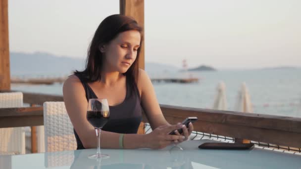 Jovem morena com copo de vinho tinto usando seu smartphone enquanto sentado no café junto ao mar ao pôr do sol . — Vídeo de Stock