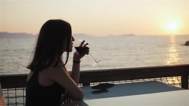 Giovane bruna con bicchiere di vino rosso che utilizza il suo smartphone mentre si siede nel caffè al mare al tramonto — Video Stock