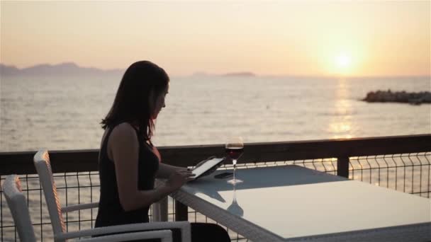 Junge Brünette mit einem Glas Rotwein mit ihrem Tablet-PC, während sie bei Sonnenuntergang im Café am Meer sitzt — Stockvideo