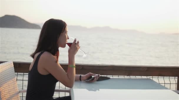 Joven morena con copa de vino tinto usando su teléfono inteligente mientras está sentada en el café junto al mar al atardecer — Vídeos de Stock