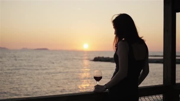 Силуэт молодой женщины, пьющей красное вино на открытой террасе, смотрящей красивый закат Вид на море — стоковое видео