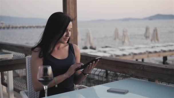 Joven morena con copa de vino tinto usando su tableta PC mientras está sentada en el café junto al mar al atardecer . — Vídeos de Stock