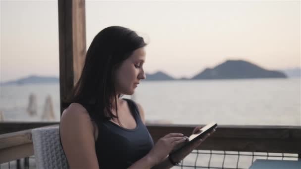 Joven morena con copa de vino tinto usando su tableta PC mientras está sentada en el café junto al mar al atardecer . — Vídeos de Stock