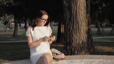 Gün batımında parkta bankta oturmuş Smartphone kullanan genç gülümseyen kadın. Güzel Avrupa kız manifatura telefon