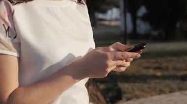 Kadının el günbatımında parkta bankta oturmuş Smartphone kullanarak kapatın. Güzel Avrupa kız manifatura telefon