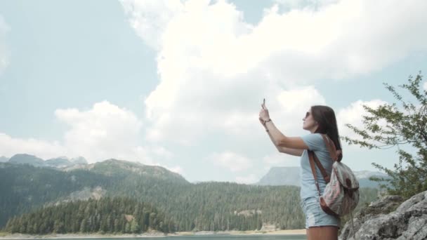 Mujer joven tomando fotos por teléfono inteligente en frente de Mountain Lake. hermosa caucásico chica pasando tiempo en un outain bosque — Vídeo de stock