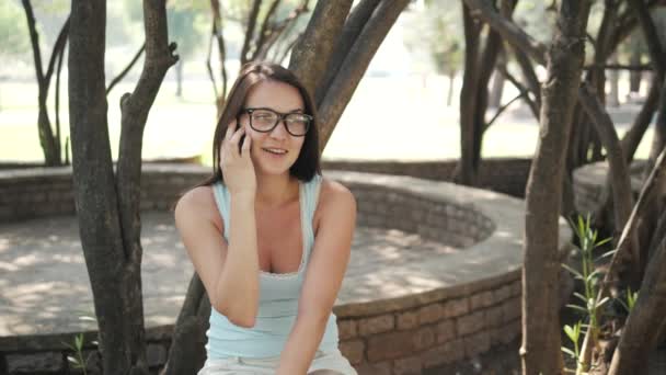 Menina alegre bonita com um smartphone sentado em um parque em um banco em um dia ensolarado — Vídeo de Stock