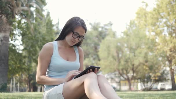 年轻美丽的女孩坐在公园的草坪上使用平板电脑, 网上购物的概念 — 图库视频影像