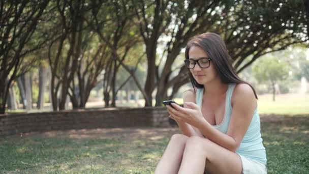 Ung vacker flicka sitter på gräsmattan i parken med telefon, On-line Shopping koncept — Stockvideo