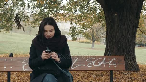 Jonge lachende vrouw met behulp van de smartphone zit op bankje in park. Mooi Europees meisje texting op telefoon — Stockvideo