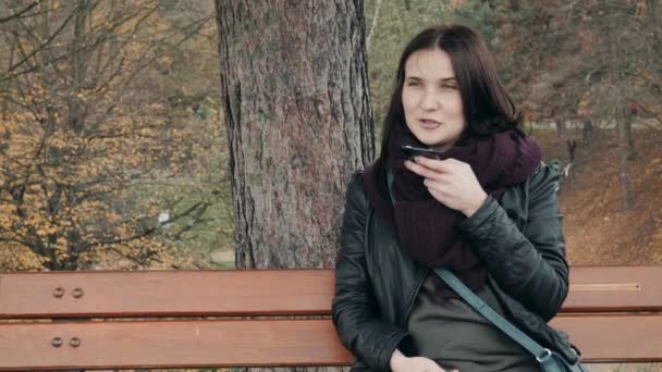 年轻的微笑的妇女使用智能手机坐在公园的长凳上。美丽的欧洲女孩在电话里做语音留言 — 图库视频影像