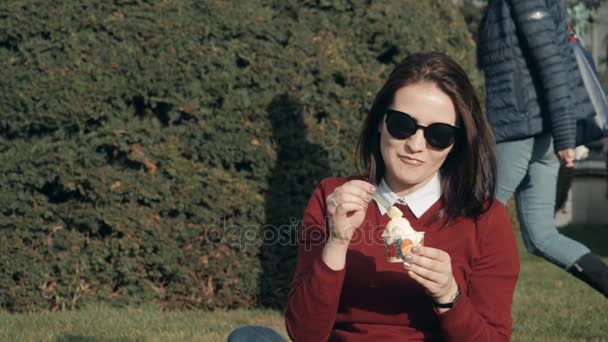 Счастливая молодая женщина наслаждается мороженым, сидя на траве в парке в солнечный день — стоковое видео