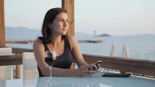 Giovane bruna con bicchiere di vino rosso che utilizza il suo smartphone mentre si siede nel caffè al mare al tramonto . Foto Stock Royalty Free