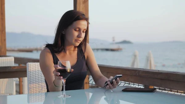Giovane bruna con bicchiere di vino rosso che utilizza il suo smartphone mentre si siede nel caffè al mare al tramonto . Immagini Stock Royalty Free