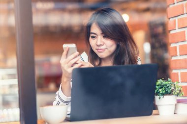 Asyalı kadın gülümseme kafede dizüstü bilgisayar ile akıllı telefon telefon kullanırken