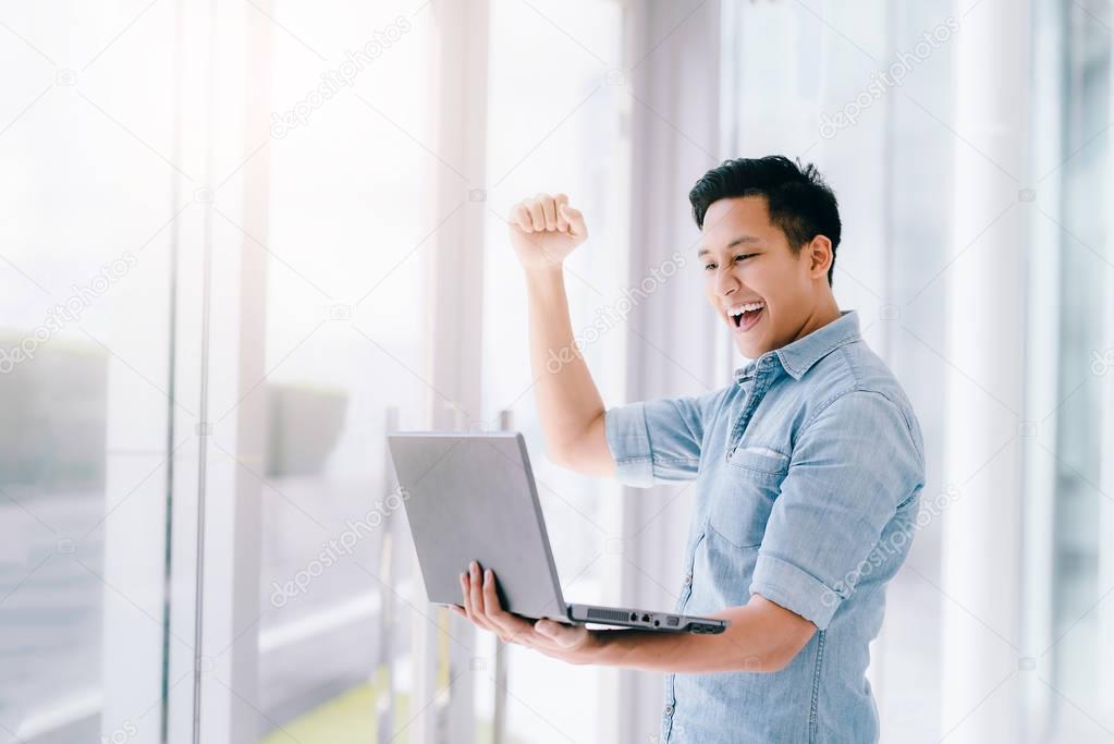 excited Asian man holding laptop and raising his arm up to celebrate success