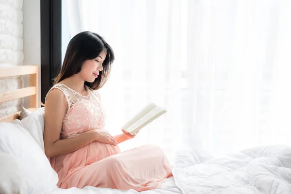 幸せ笑顔の寝室に注意を彼女の腹に触れる中で 本を読んでアジア妊婦 — ストック写真