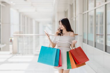 Güzel genç mutlu Asyalı kadın ile çarşıda alışveriş yaparken smartphone kullanarak renkli alışveriş çantası