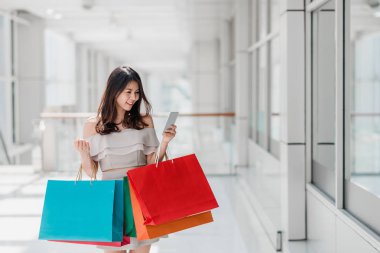 Güzel genç mutlu Asyalı kadın ile çarşıda alışveriş yaparken smartphone kullanarak renkli alışveriş çantası