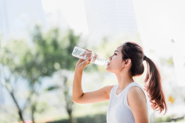Genç Asyalı kadın içme suyu su parkta koşu sonra şişe bardak.