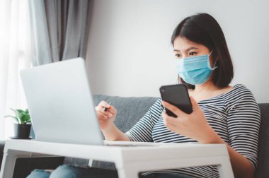 Karantina sırasında evde çalışan Asyalı kadın dizüstü bilgisayar ve akıllı telefon kullanarak yüz maskesi takıyor