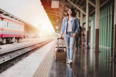 Güzel gülümseyen Asyalı turist kadın seyahat sırasında tren istasyonundaki platformda bavulla yürüyor.