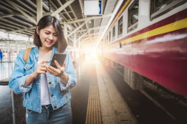 Akıllı telefon kullanan mutlu Asyalı kadın turist tren istasyonunda yön arıyor.