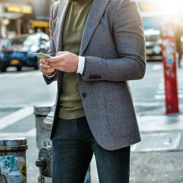 Stile street fashion maschile. Giacca classica a due bottoni felpa verde e jeans grigi. Uomo elegante a piedi sulla strada della città e sms sul telefono cellulare . — Foto Stock