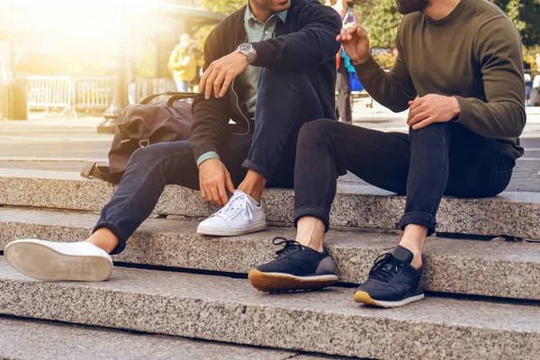 Lifestyle photo of two male friends sitting on the steps in city street and talking wearing casual street style clothes and sneakers. — Stock Photo, Image