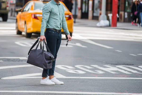 Homem elegante andando na rua da cidade passarela vestindo roupas casuais com jeans t-shite formiga e segurando saco de viagem e óculos de sol nas mãos — Fotografia de Stock